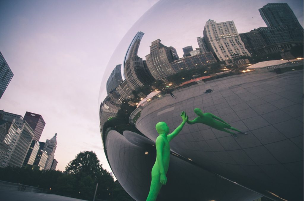 Artistic photo of a person fully covered in green fabric is touching a big mirror ball, in some modern city centre