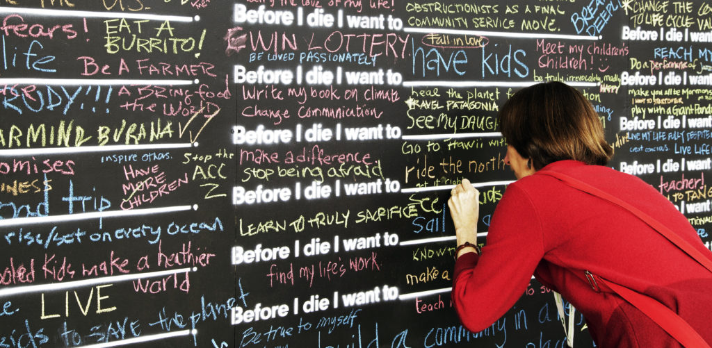 A woman in red pullover is writing on the before I die wall